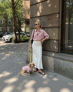 Woman in pink shirt and white skirt standing on a sidewalk with a dog in a pink outfit.