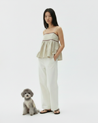 Woman in a strapless top and white pants standing next to a small dog on a plain background