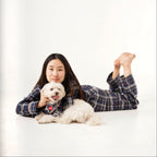 Person lying on a white floor with a small white dog, both wearing plaid shirts.