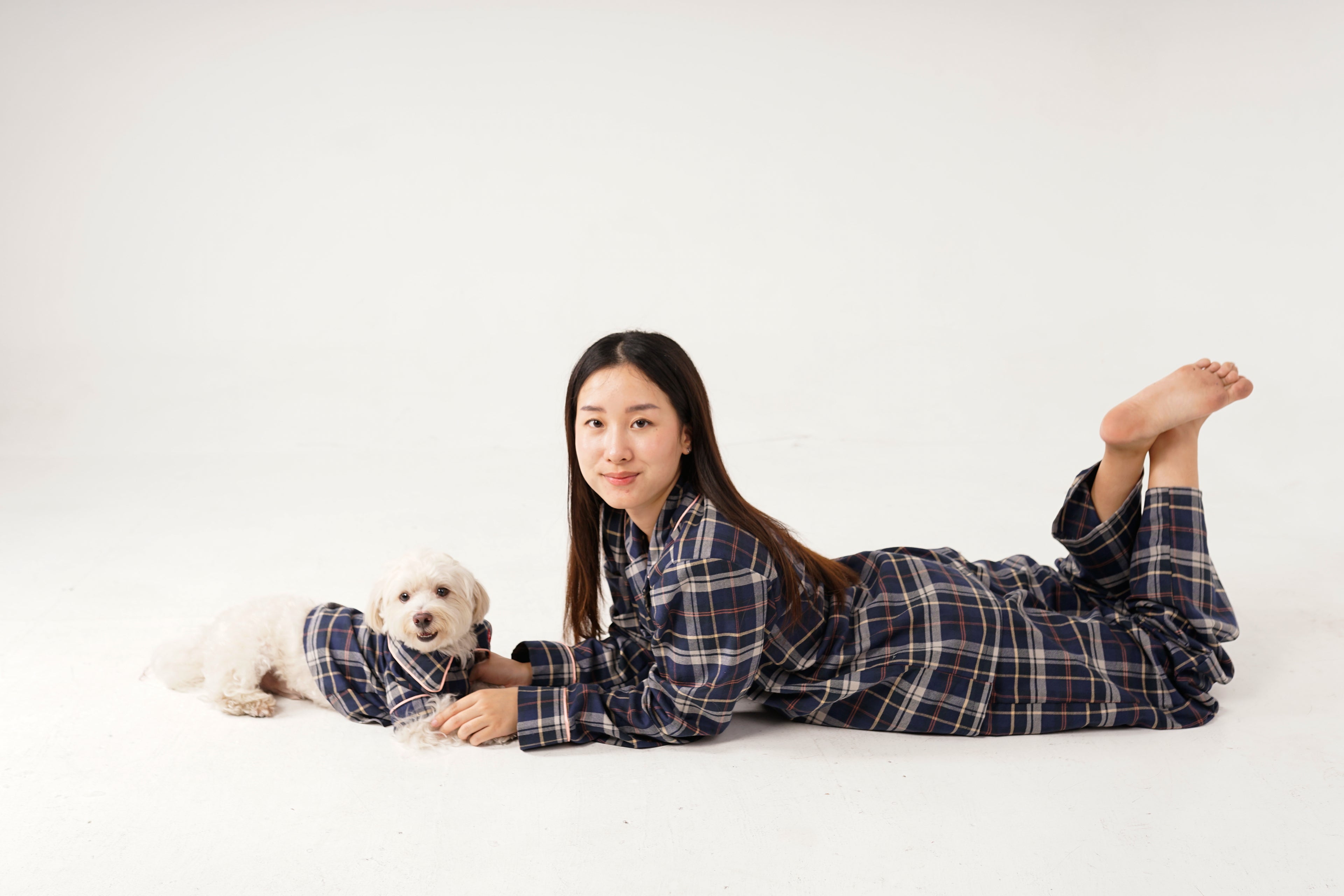 Woman in plaid pajamas lying on a white floor with a small dog next to her.