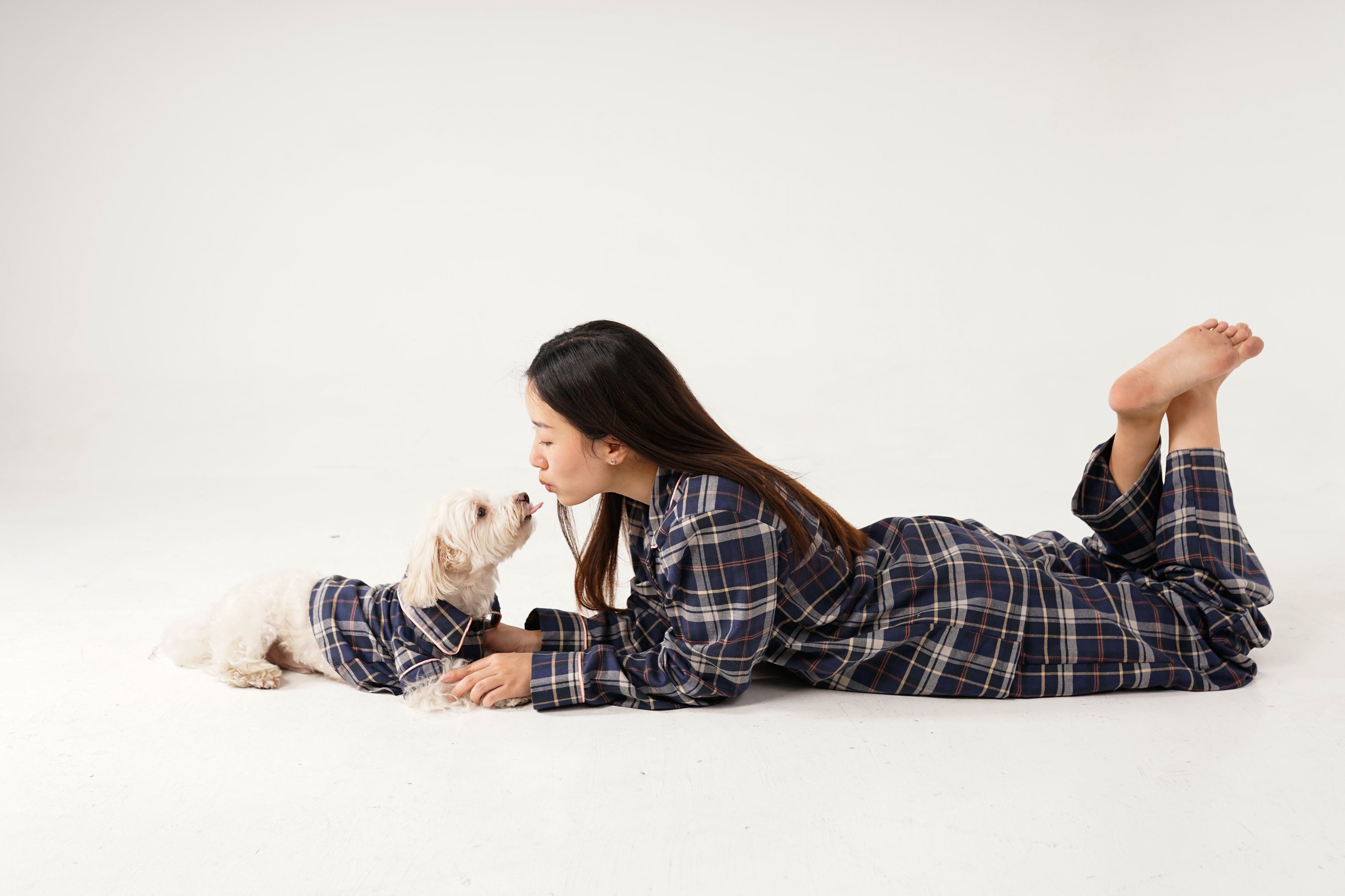 Woman in plaid pajamas lying on a white floor with a small white dog.