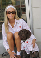 Woman in a striped shirt and sunglasses with a dog wearing a matching outfit, sitting on a sidewalk.