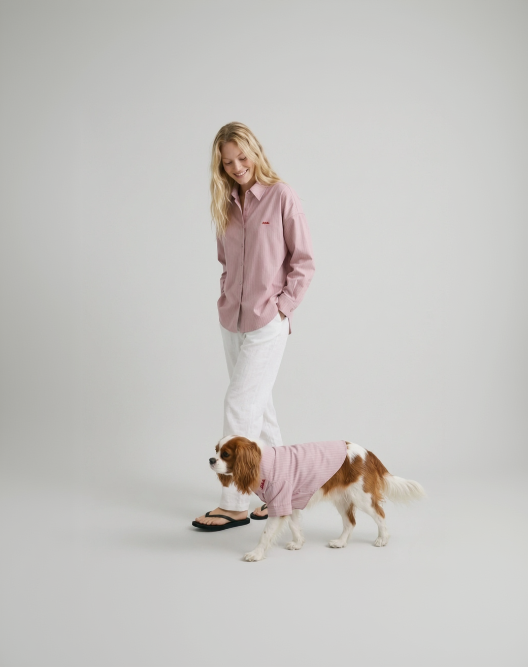 Woman in pink shirt and white pants standing next to a dog wearing a pink sweater on a plain background