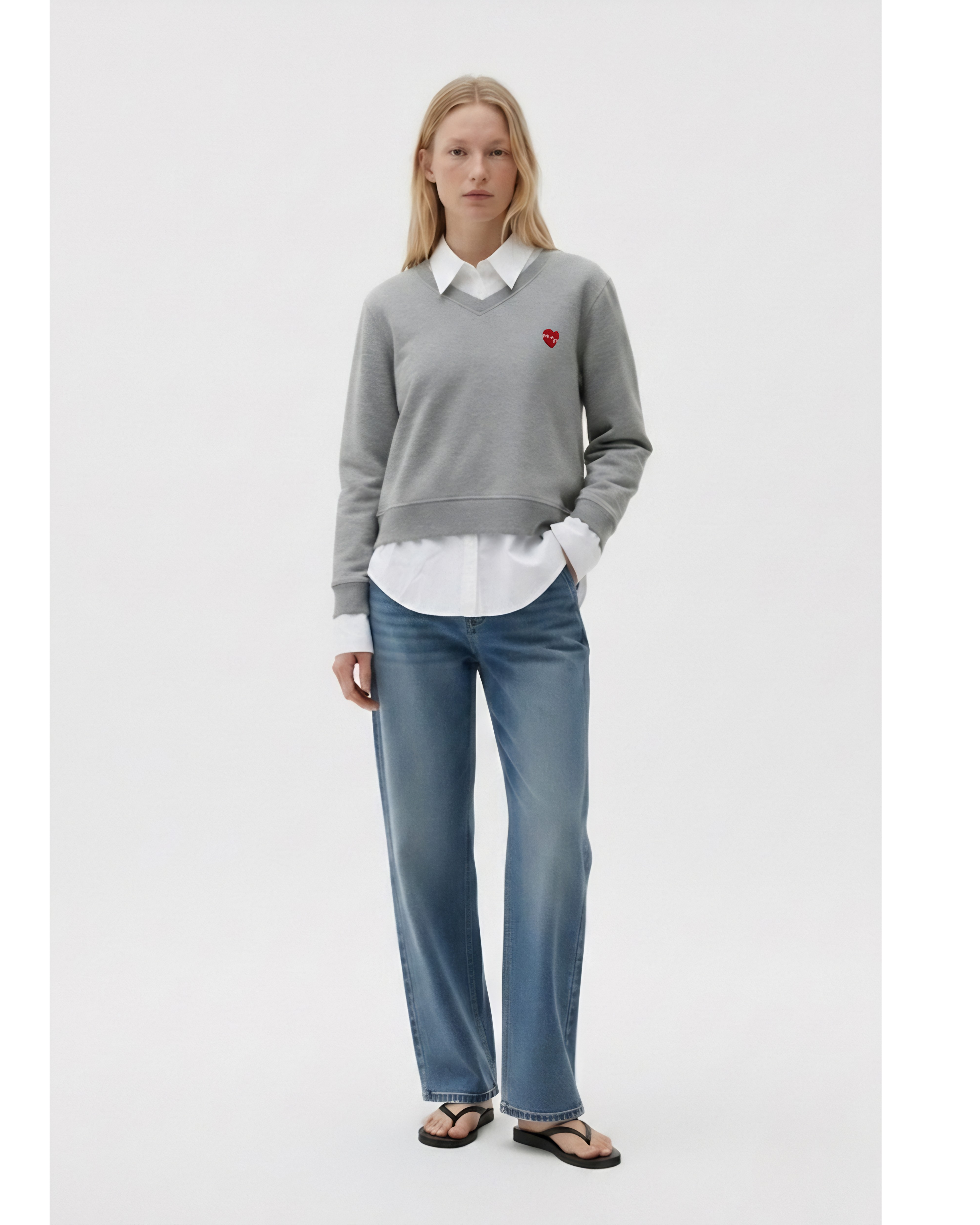 Woman wearing a gray sweater with a red heart logo, white shirt, and blue jeans on a white background